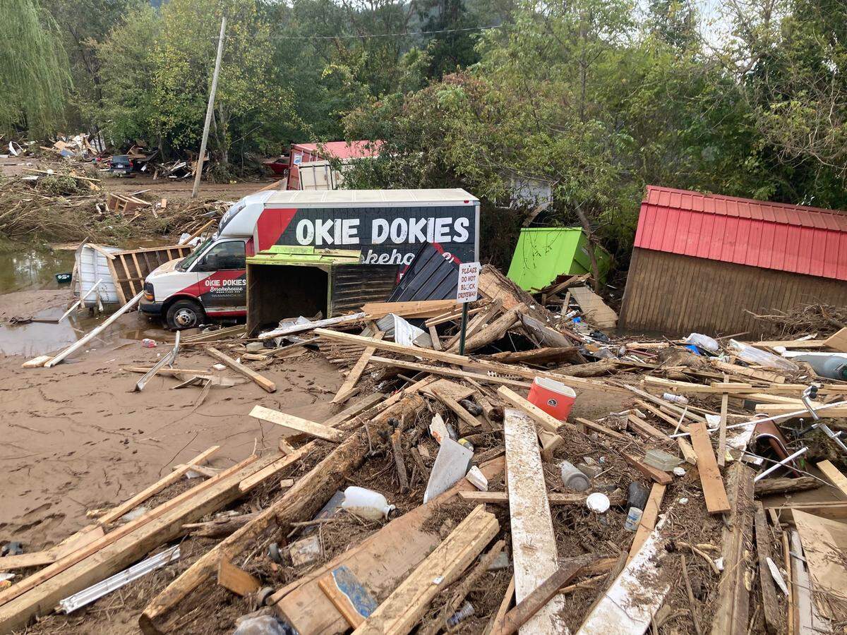 The aftermath of a major flood. In the center, an Okie Dokies Smokehouse delivery van is stuck at an angle in muddy water. The entire area is covered in a thick layer of mud and a huge, tangled pile of debris from destroyed structures, including wooden beams and pieces of sheds.
