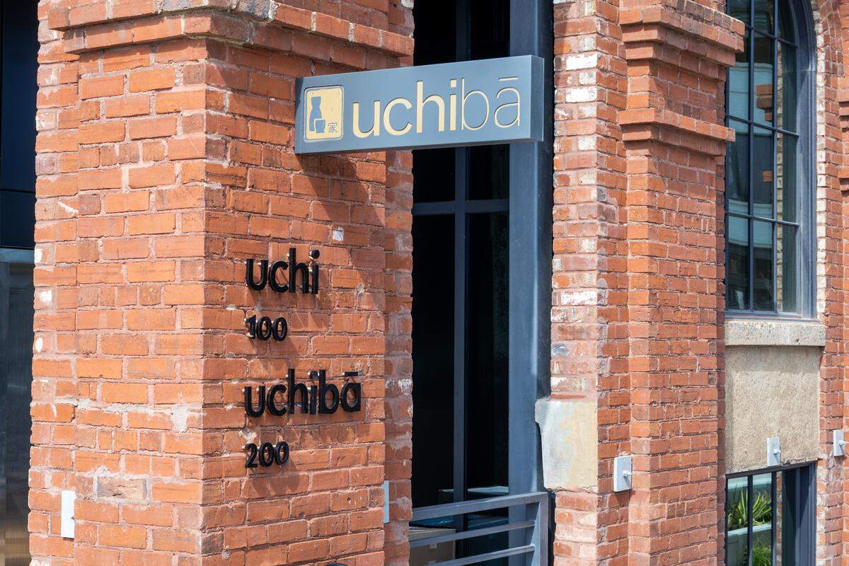 A close-up of a red brick pillar featuring the Uchi and Uchiba logos in black lettering. Above them, a grey rectangular sign with gold lettering and a sake bottle icon also reads “uchibā”.