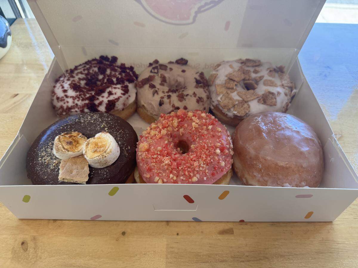 A top-down view of a box containing six unique doughnuts. The top row, from left to right, shows a red velvet doughnut with cream cheese frosting and red velvet crumbles, a chocolate-frosted doughnut with milk and dark chocolate chips, and a vanilla-glazed doughnut topped with Cinnamon Toast Crunch cereal. The bottom row, from left to right, shows a chocolate-frosted doughnut with graham cracker pieces and toasted marshmallows, a strawberry-frosted doughnut with strawberry shortcake crumbles, and a classic glazed doughnut.