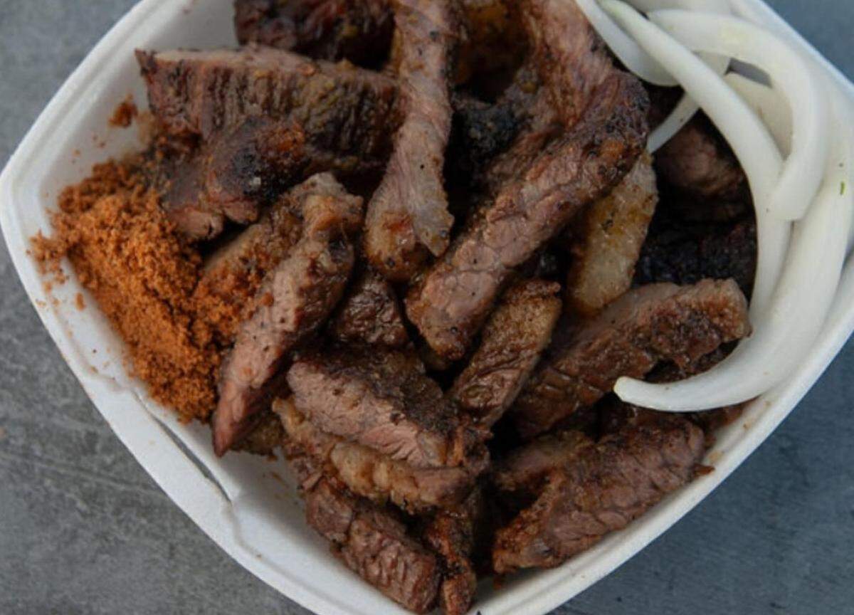 A top-down, close-up shot of a serving of suya in a white disposable container. The dish consists of numerous strips of grilled, charred beef that are piled in the center of the container. A generous pile of orange-brown yaji (spicy peanut-based seasoning) is tucked into the left corner of the container. Several thick slices of raw white onion are placed on the right side of the meat.