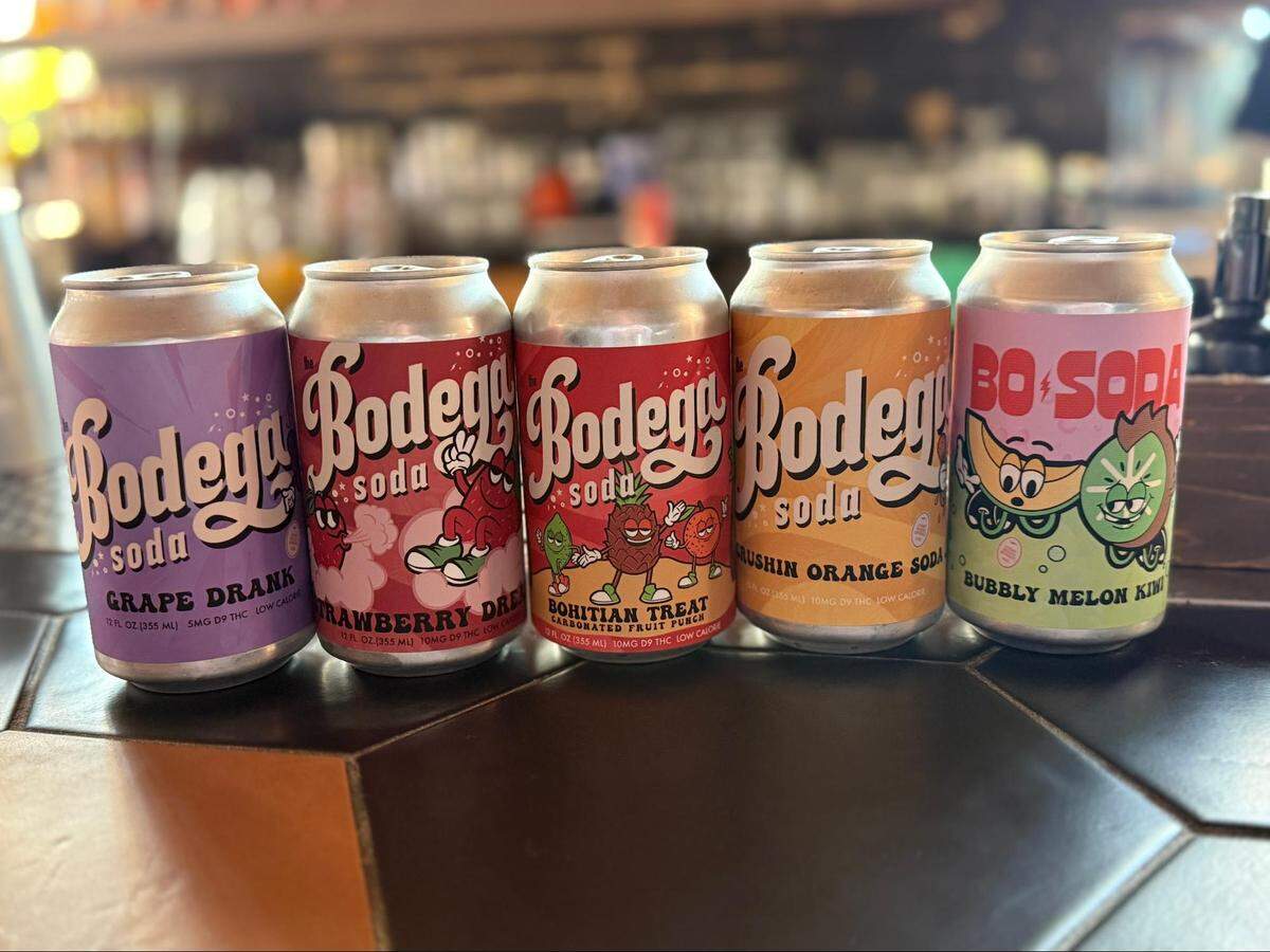 A close-up, eye-level shot of five colorful aluminum soda cans lined up on a dark tiled bar. The background is a blurred bar with shelves of bottles. From left to right, the cans are: “Bodega soda Grape Drank,” “Bodega soda Strawberry Dream,” “Bodega soda Rohitan Treat” fruit punch, “Bodega soda Crushin Orange Soda,” and “Bo Soda Bubbly Melon King.”
