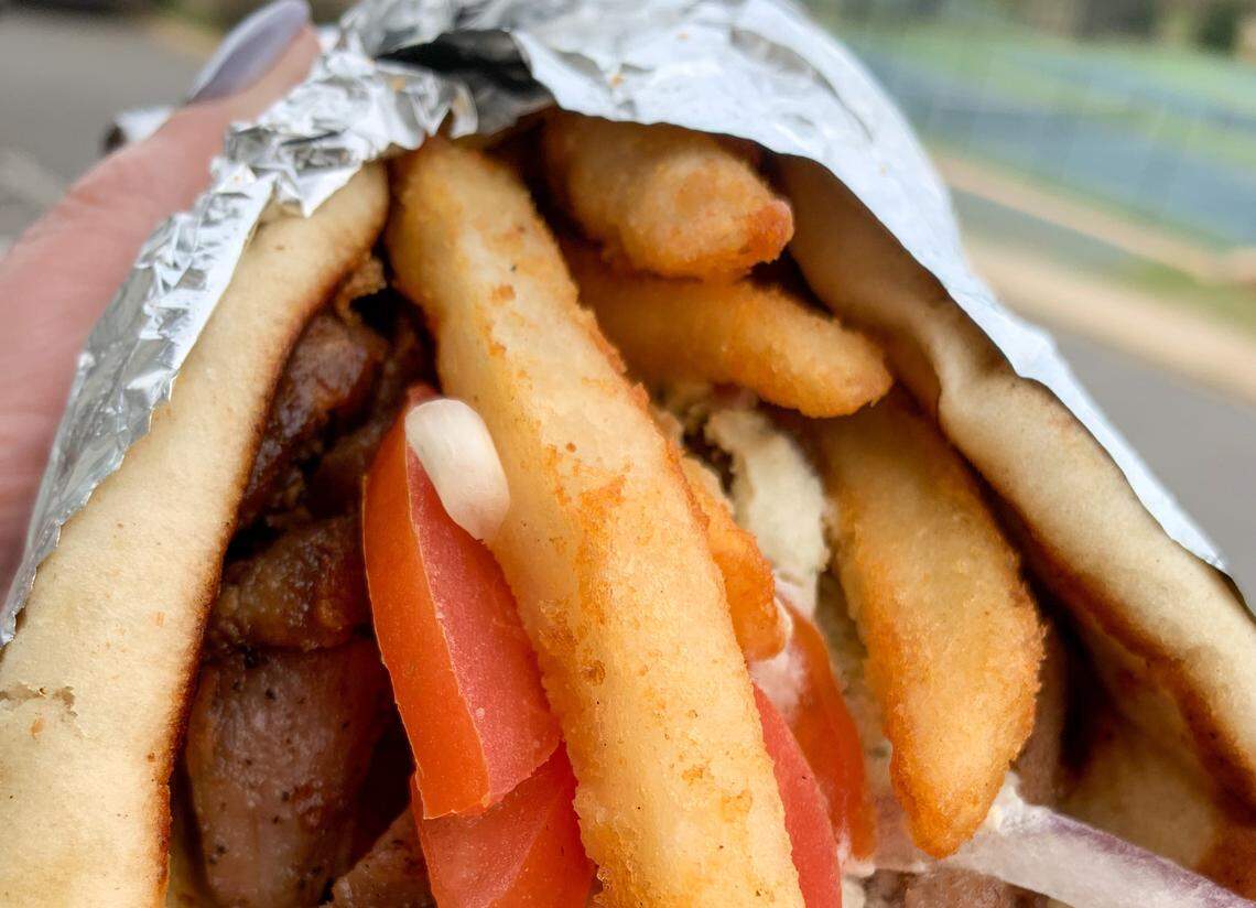 An extreme close-up of a gyro sandwich held in a person’s hand. The pita is packed with traditional fillings like gyro meat and tomato, as well as a handful of golden french fries. The sandwich is partially wrapped in aluminum foil.