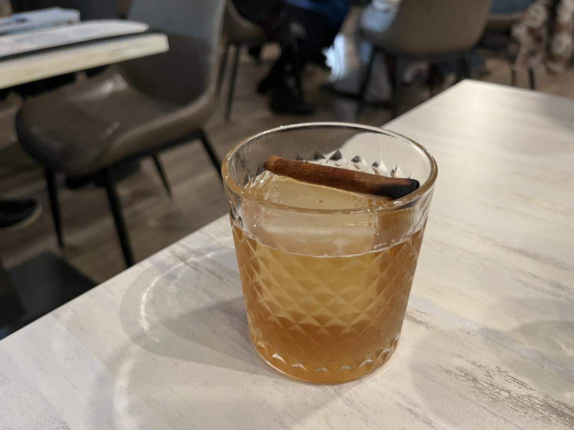 A close-up shot of a cocktail on a light-colored, wood-grain table. The drink, an amber-colored liquid, is in a textured, diamond-patterned rocks glass with a large square ice cube, and is garnished with a single cinnamon stick resting on top. The background shows a blurred view of the restaurant interior, including a chair.