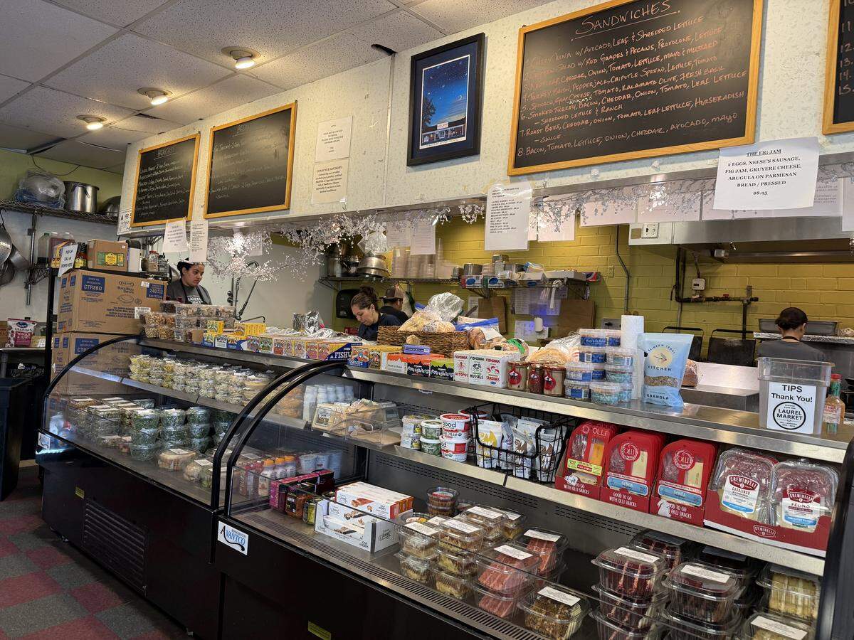 The bustling interior of a deli, featuring a long, curved glass display case filled with pre-packaged sandwiches, salads, and other grab-and-go items. Behind the counter, employees are working, with large chalkboard menus listing sandwiches hanging on the wall above them.