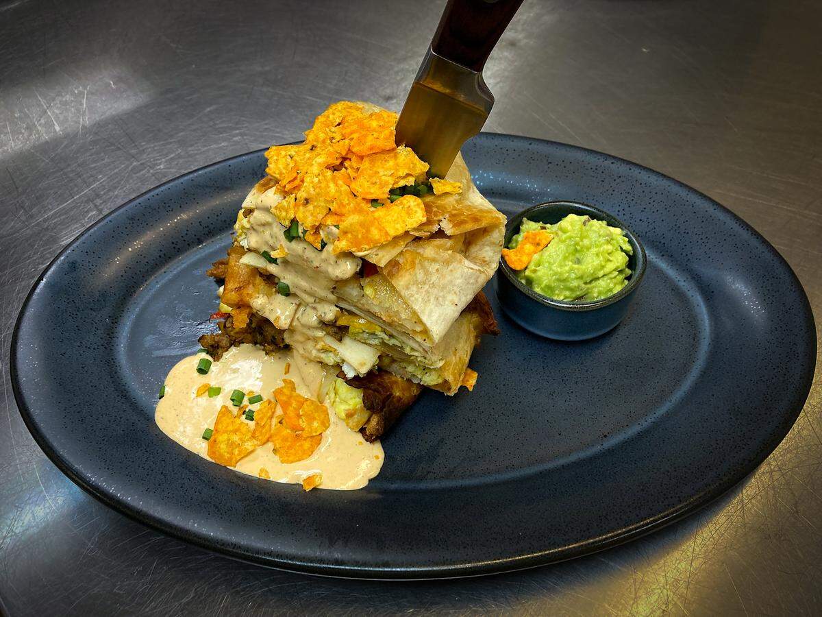 On a large, dark grey plate in a restaurant kitchen, a deconstructed crunch wrap is presented as a tall stack of layered tortillas, meat, and eggs. The dish is finished with a creamy chipotle-style sauce, crushed chips, and a large knife stuck straight down through the center. A small bowl of guacamole accompanies the dish.