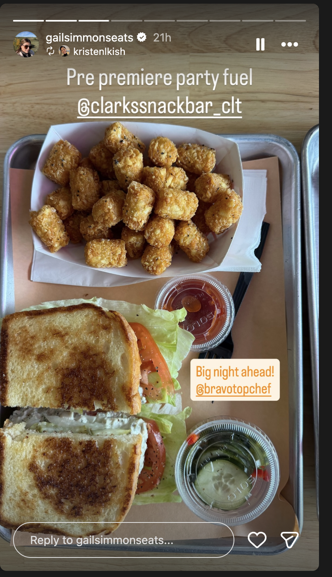 An overhead shot of a metal food tray from a social media story by @gailsimmonseats, featuring a meal from @clarkssnackbar_clt. The meal consists of a large portion of golden-brown tater tots in a white paper boat, a toasted sandwich cut in half, filled with what appears to be tuna or chicken salad, accompanied by fresh lettuce and tomato slices, and two small plastic containers: one holding a red sauce (likely ketchup or jam) and another with sliced pickles.