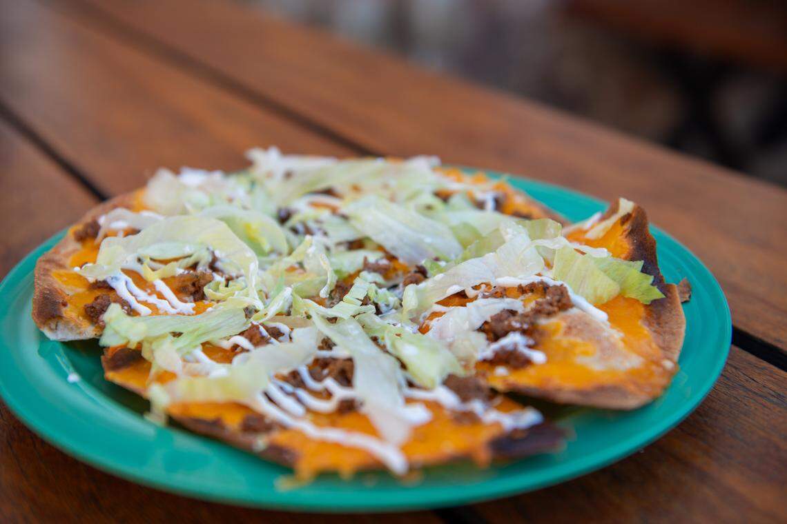 An angled, close-up shot of a Mexican pizza-style dish on a bright turquoise-green plate. The dish is covered in melted cheese, seasoned ground meat, shredded lettuce, and a zigzag drizzle of white cream. The plate is set on a dark wooden table.