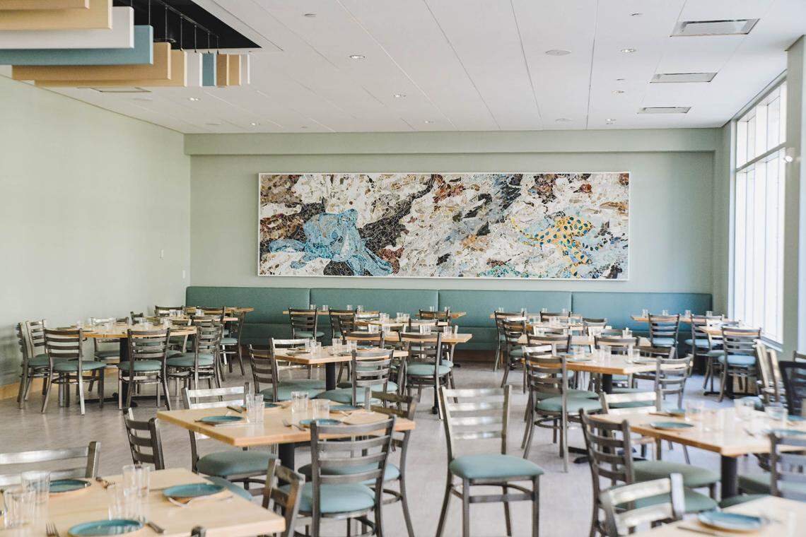 A wide, brightly lit dining room featuring light wood tables and metal chairs with teal cushions. In the background, a long teal banquette sits against a light green wall, beneath a large horizontal mosaic artwork depicting marine life, including an octopus and a sea turtle. The room has high ceilings with white acoustic tiles and large windows on the right side that fill the space with natural light. Several tables are set with teal plates and clear glassware, ready for service.
