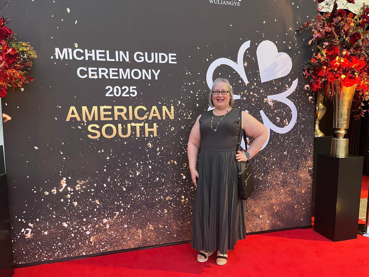 A smiling person with short gray hair and glasses is standing on a red carpet in front of a dark backdrop for the “MICHELIN GUIDE CEREMONY 2025 AMERICAN SOUTH.” Ther person is wearing a dark gray pleated sleeveless dress and black sandals, and holding a black handbag with a right hand on a hip. The backdrop is black with sparkling gold confetti effects and features a large white Michelin Guide logo with a four-leaf clover design. Tall vases overflowing with dark red flowers flank the backdrop.