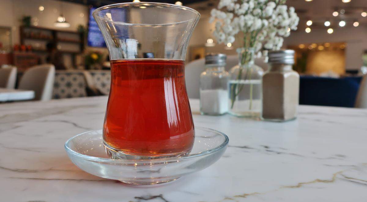 A close-up of a traditional tulip-shaped glass filled with Turkish tea, sitting on a matching glass saucer on a white marble table. The background shows the blurred interior of the cafe.