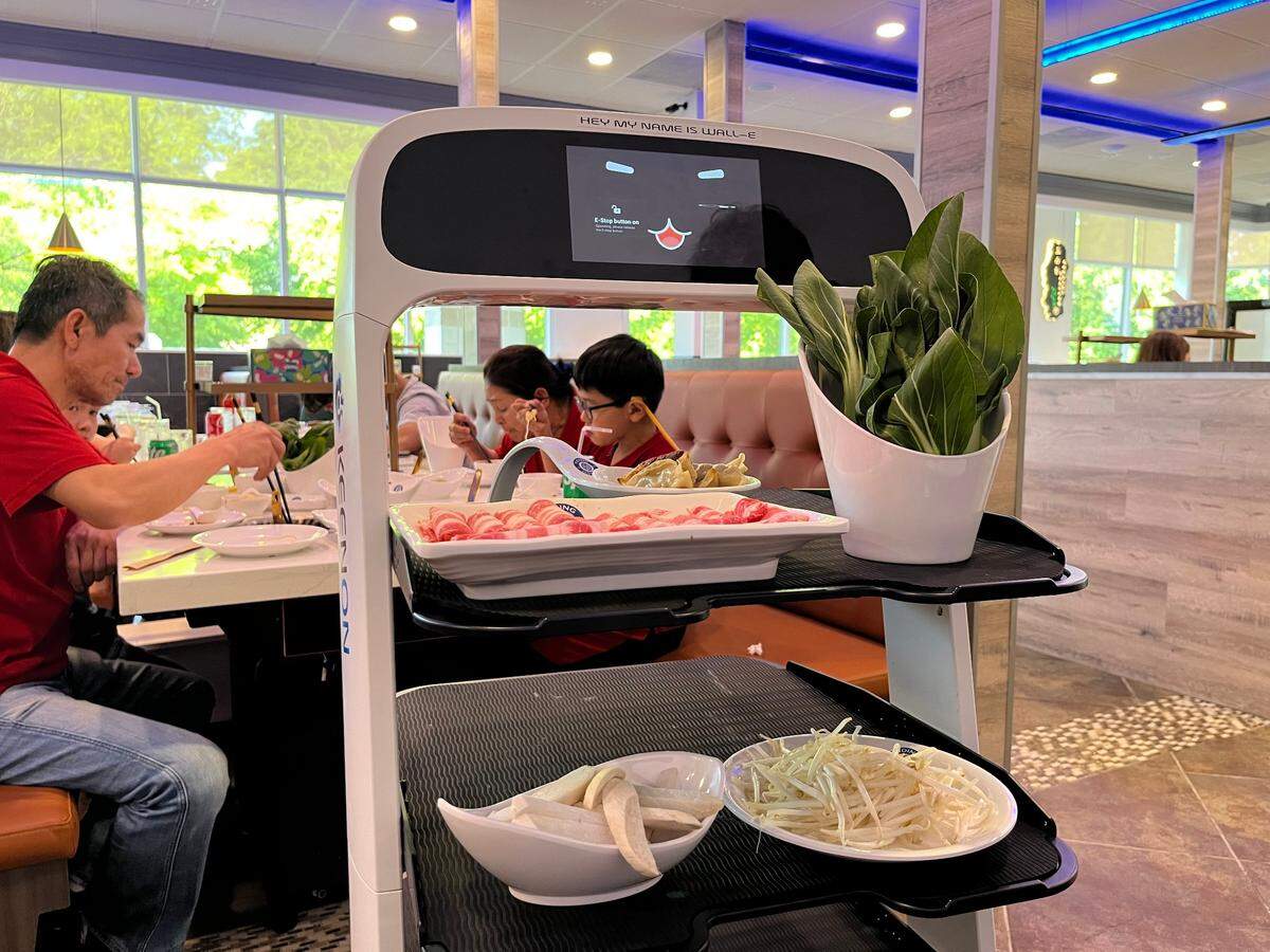 A white, automated food delivery robot is paused in the aisle of a modern restaurant. Its screen displays a digital face and its name, “WALL-E.” The robot’s shelves are filled with fresh hot pot ingredients, including a platter of thinly sliced meat and bowls of vegetables, while patrons dine at tables in the background.