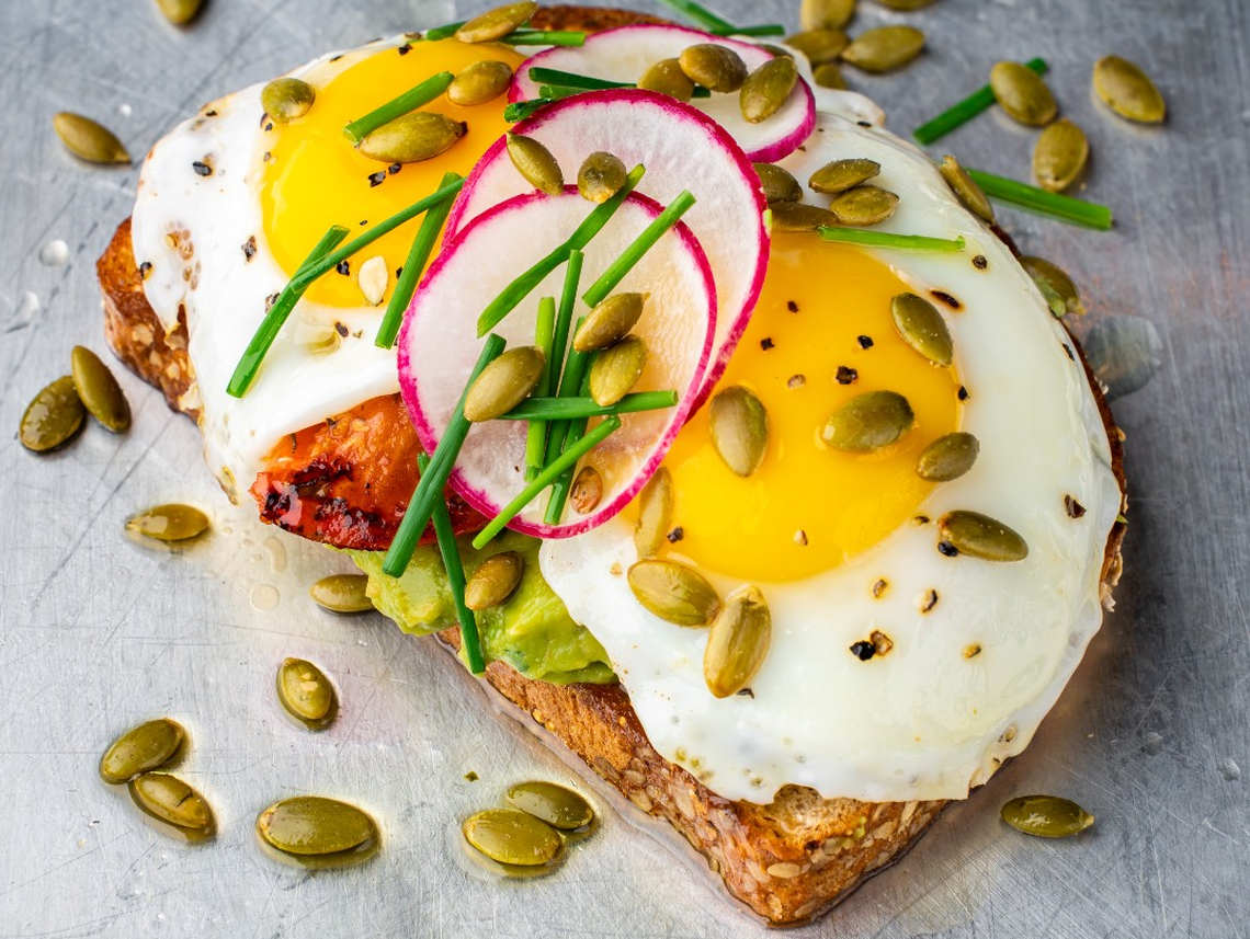 A high-angle, close-up shot of a single slice of avocado toast on a metallic surface. The toast is topped with mashed avocado, two sunny-side-up eggs, and garnished with thin slices of radish, chopped chives, and pumpkin seeds.