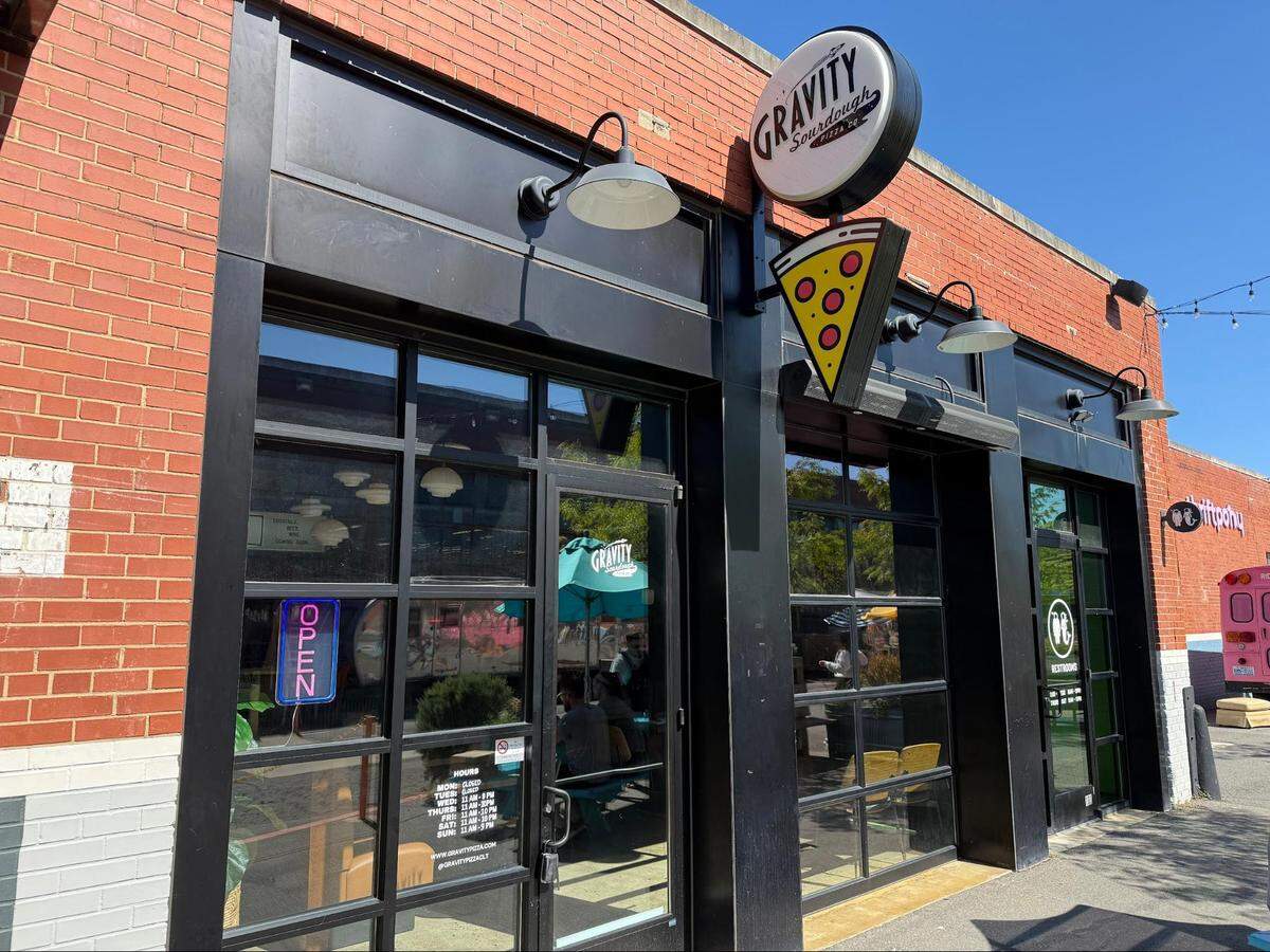 A straight-on, eye-level shot of the exterior of a “Gravity Sourdough Pizza” shop on a sunny day. The restaurant is set in a one-story, red-brick building with large, black-framed glass windows and doors. A round white sign with the restaurant’s name hangs above, along with a separate, illuminated sign in the shape of a pizza slice. A blue and red neon “OPEN” sign is visible in the window of the left-hand door.
