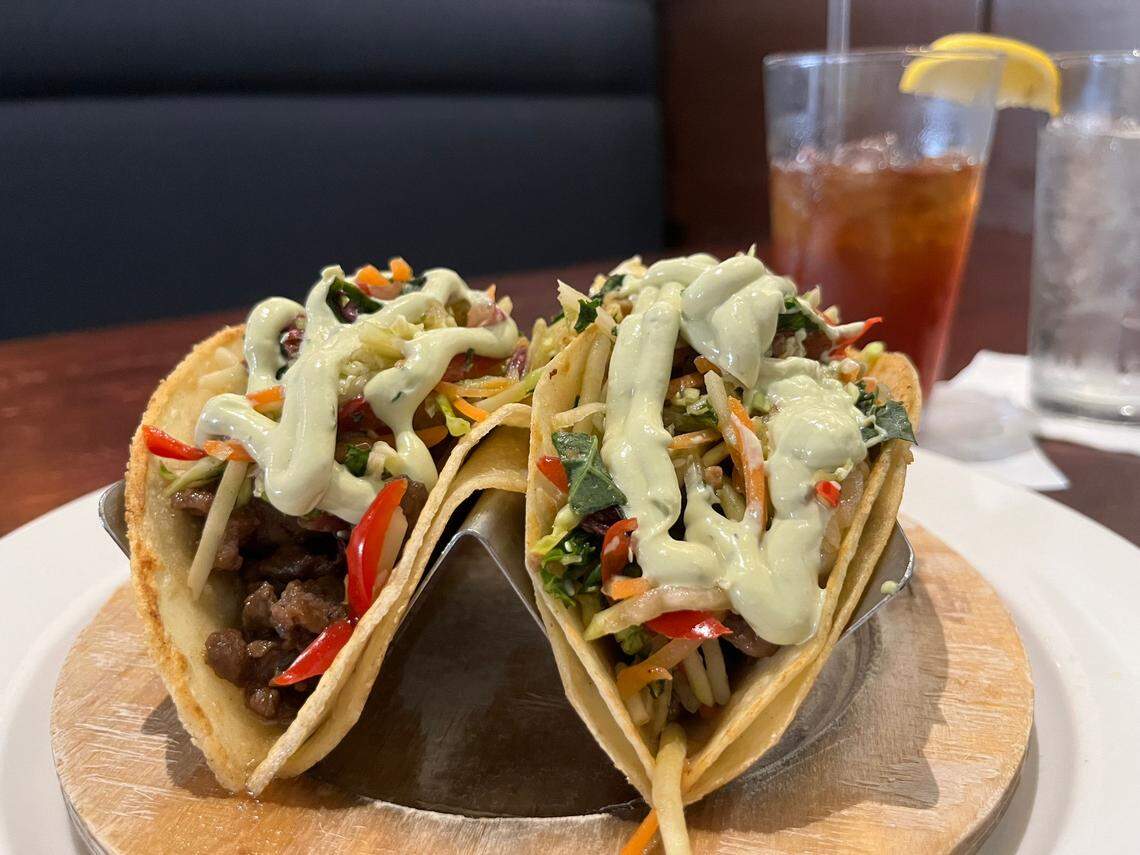 A pair of tacos is presented on a wooden stand inside a white plate. The tacos have hard, crispy shells filled with what appears to be ground meat or seasoned beef, a colorful mix of shredded vegetables like carrots and red bell peppers, and a drizzle of a light green creamy sauce. In the blurred background, a glass of iced tea with a lemon wedge and a glass of water are visible. The setting appears to be a restaurant with a dark booth in the back.