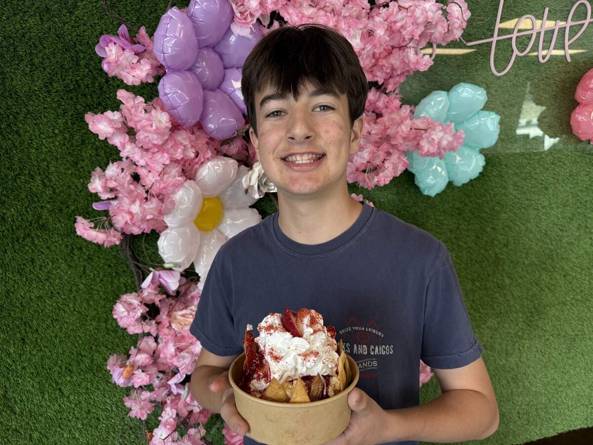 A teenager smiles and holds a dessert bowl topped with whipped cream and strawberries in front of a pink flower wall.