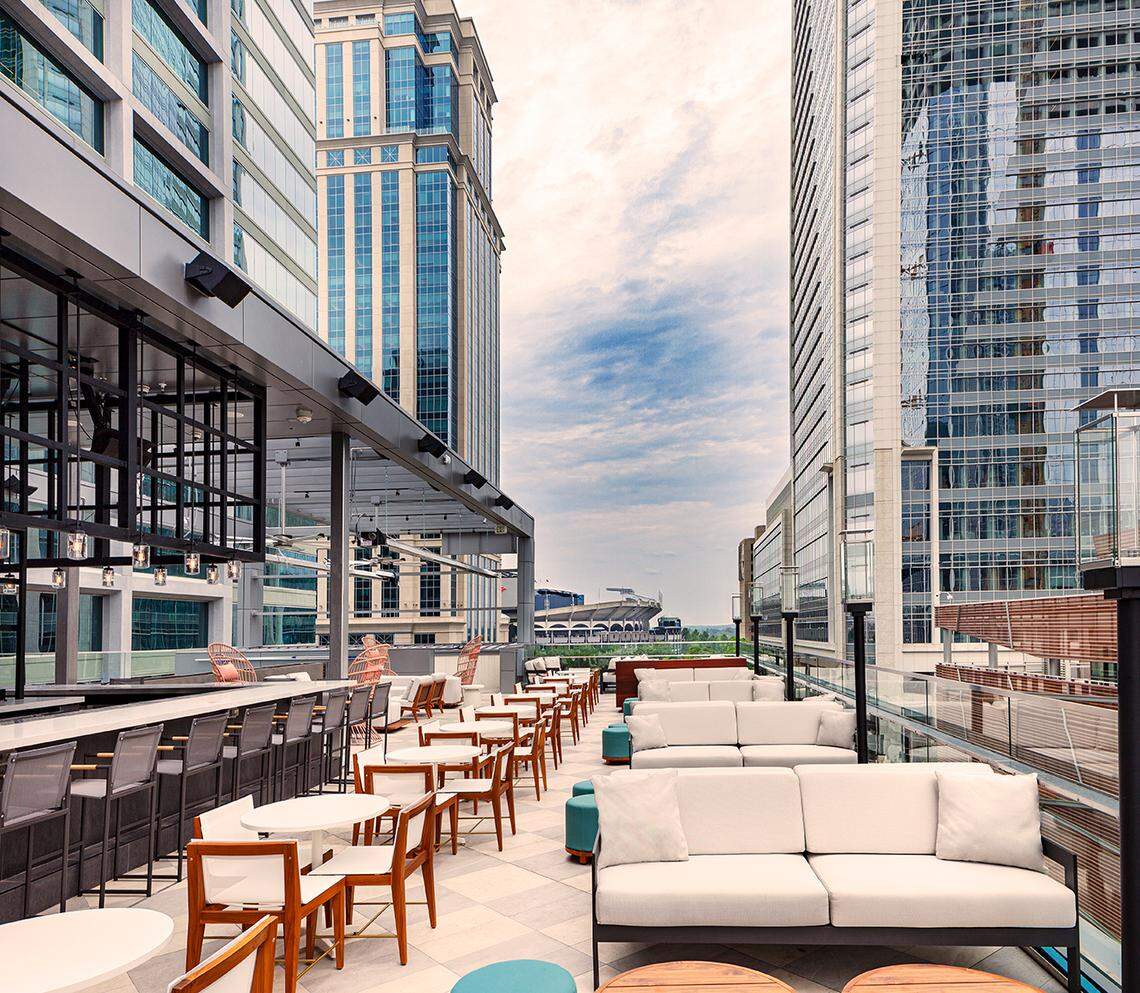 This daytime image shows a chic rooftop restaurant or bar set between tall city buildings with a mix of seating areas. The space features a long bar with stools, white tables with wooden chairs, and comfortable white outdoor couches, all with a view of a nearby sports stadium.