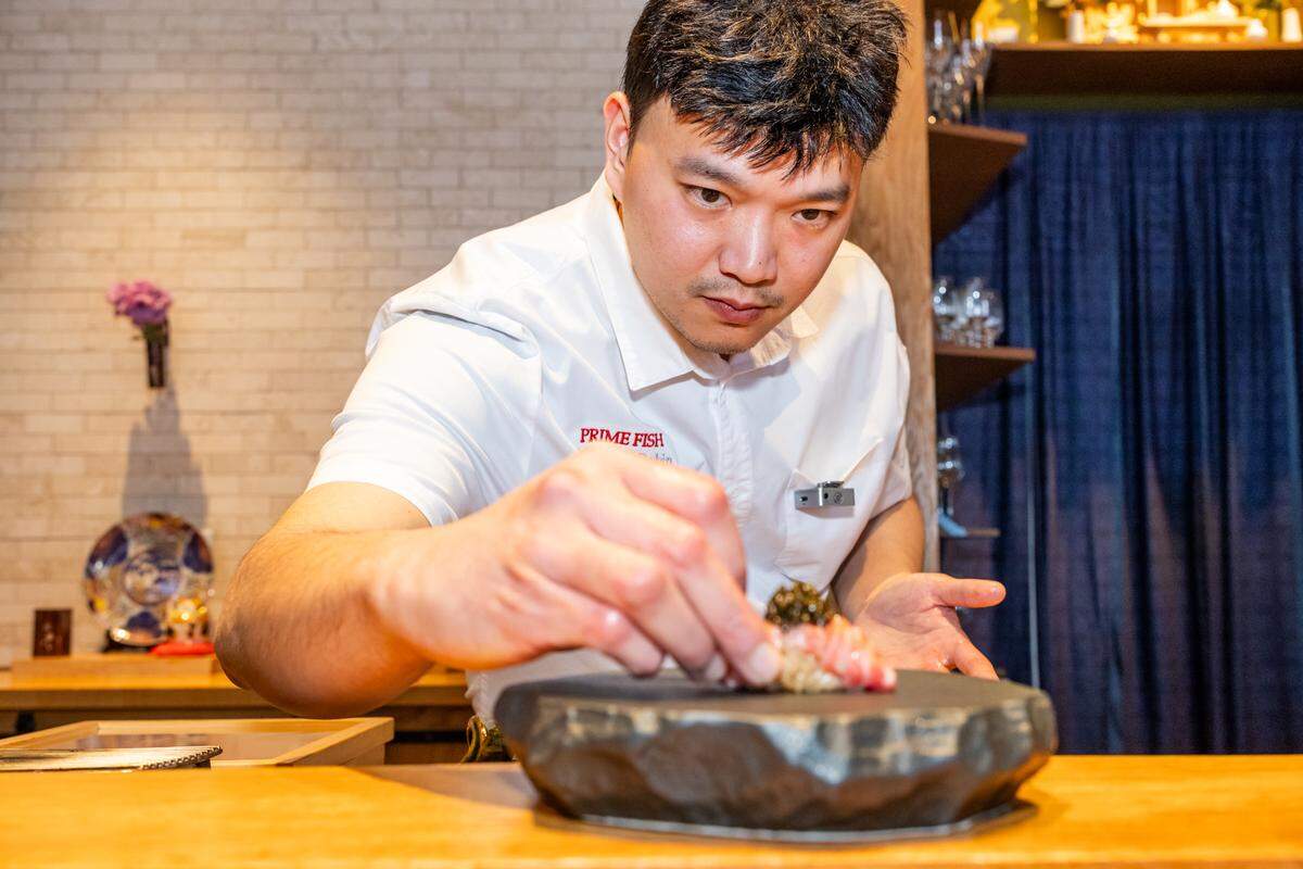 A medium-shot, eye-level photograph shows a chef with short black hair, wearing a white short-sleeved button-down shirt with “PRIME FISH” and “Chef Robin” embroidered in red on the right chest, leaning forward over a light-colored wooden counter and focusing intently to delicately place a piece of sushi onto a dark, textured, stone-like plate.