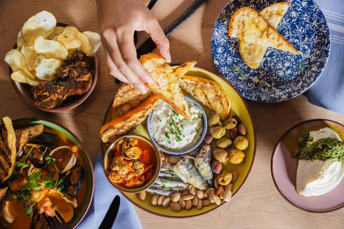 A high-angle shot captures a vibrant spread of dishes on a wooden table, with a person’s hand reaching for a piece of toast. The central dish is a large round platter with a yellow rim, holding a bowl of dip topped with chives, surrounded by toasted bread, a pile of green olives, several small fish (sardines), and roasted peanuts. To the left, a dark brown bowl contains what appears to be braised meat with potato chips. Below it, a green bowl is filled with a seafood stew featuring mussels, clams, and bread. To the top right, a blue and white patterned plate holds more toasted bread. On the bottom right, a light pink plate has a dollop of white cream cheese or burrata topped with green pesto.