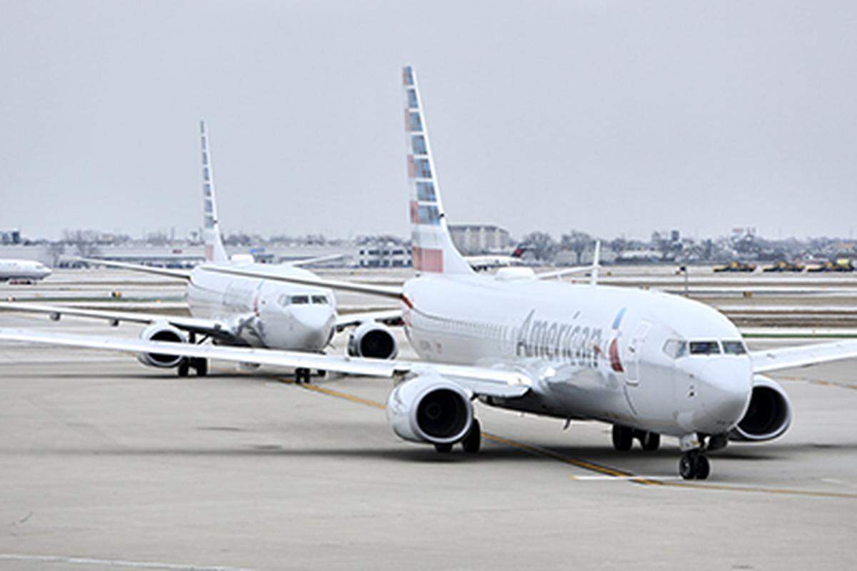 Two white American Airlines Boeing 737 aircraft are on the tarmac, with one airplane following closely behind the other. The aircraft are visible under a gray, overcast sky, suggesting a cold or winter day at the airport.