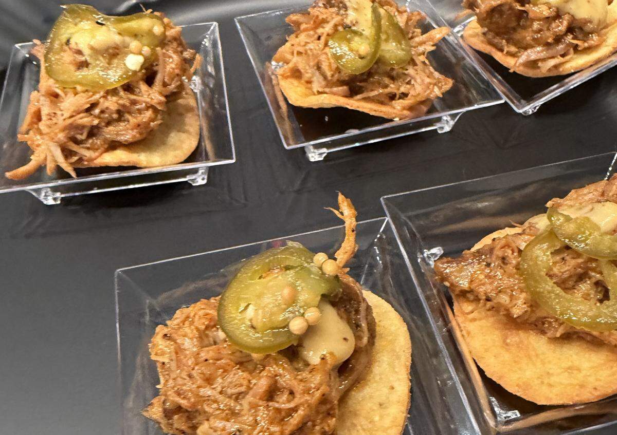 Appetizer portions of a signature dish appear in small, clear plastic square trays, each holding a miniature tostada. The tostada is topped with a generous portion of tender pulled pork, a dollop of creamy sauce, and a single pickled jalapeño slice for a kick of heat.