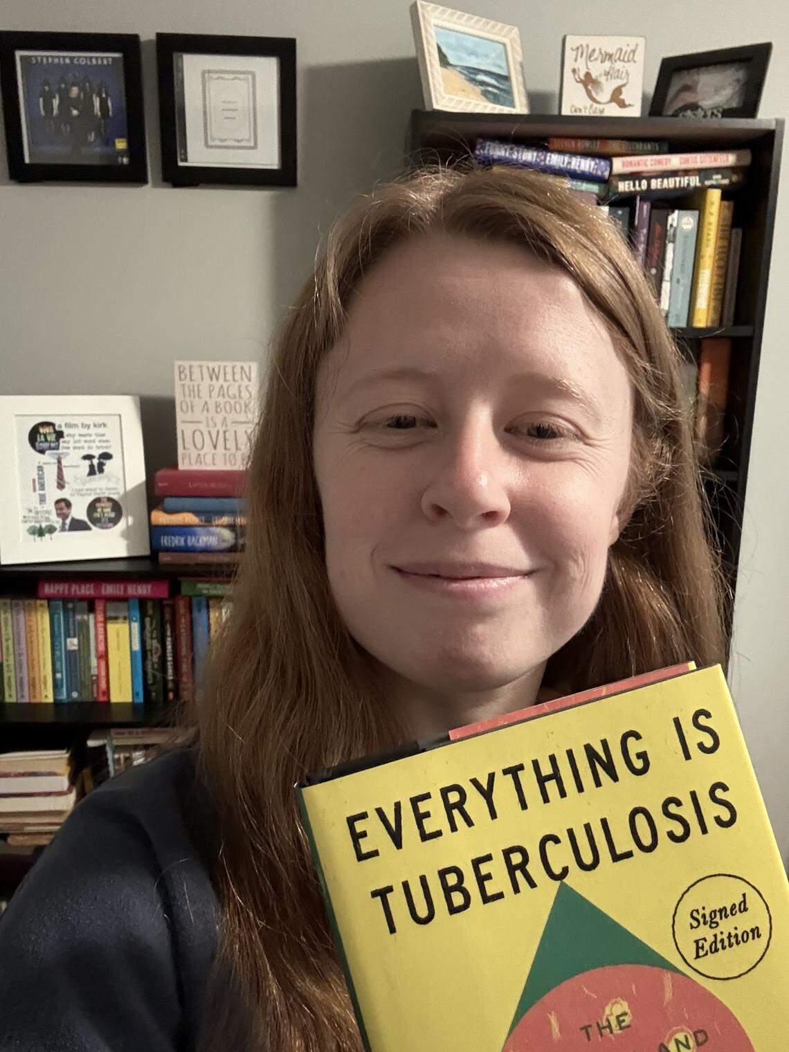 A smiling person with long, reddish hair is holding a yellow book titled “Everything Is Tuberculosis” toward the camera. They are standing in front of a densely packed bookshelf containing many books and framed pictures, including one labeled “Stephen Colbert.