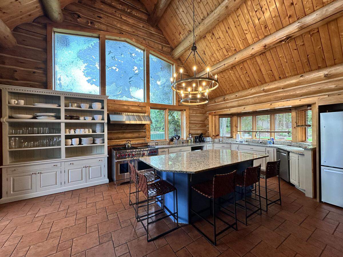 A wide view of a luxurious kitchen inside a log cabin. The room has terracotta tile floors and a very high, wood-paneled vaulted ceiling. In the center is a large, dark-blue island with a granite countertop and four bar stools. Above it hangs a large circular chandelier. Along the back wall are stainless steel appliances, a large grey hutch, and a prominent window with a frosted tree design.