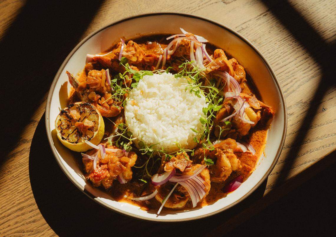 A top-down, close-up shot of Camarones Kora served in a shallow white bowl. A mound of white rice sits in the center, surrounded by large shrimp coated in a rich, reddish-orange sauce. The dish is garnished with thin slivers of red onion and fresh microgreens, accompanied by a charred lemon half. Deep shadows and bright sunlight stretch across the wooden tabletop.