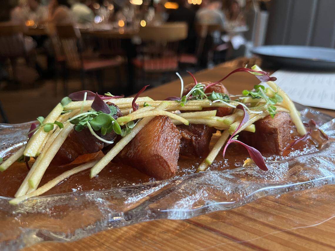A close-up, eye-level shot of an artfully plated appetizer. The dish features several cubes of cooked pork belly with a rich, glossy glaze, served on a clear, textured glass plate. The pork belly is topped with a garnish of thin apple matchsticks, fresh green and purple microgreens, and small red garnishes. The plate sits on a wooden table, and the background is a bustling, out-of-focus restaurant dining room with seated patrons.