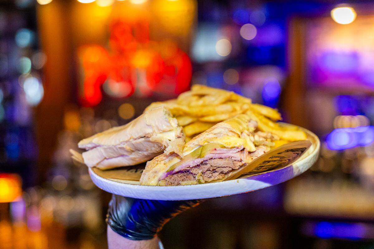 A close-up shot of a gloved hand holding up a plate with a Cuban sandwich and french fries. The sandwich is cut in half, revealing layers of pork, ham, melted cheese, and pickles. The background is the blurred, colorful, and warmly-lit interior of a bar or restaurant.
