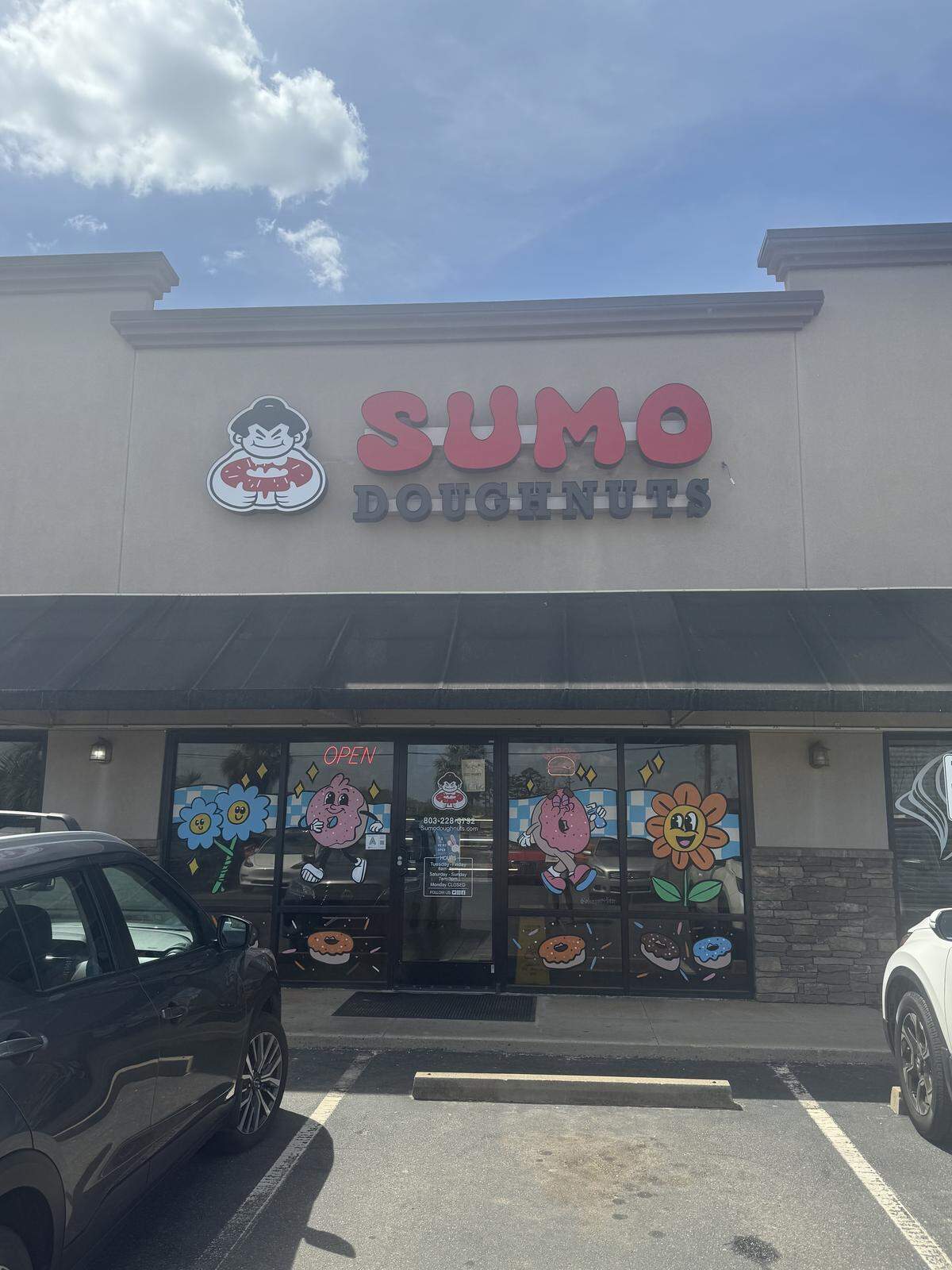The exterior of Sumo Doughnuts, a storefront in a strip mall. The sign features a red “SUMO” in a bold, rounded font and “DOUGHNUTS” in gray. To the left is a logo of a sumo wrestler holding a large doughnut. The windows are decorated with colorful, retro-style cartoon illustrations of smiling flowers and anthropomorphic doughnuts.