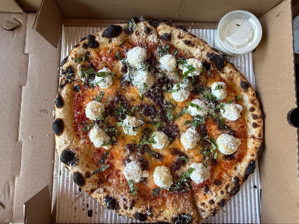 An overhead, close-up shot of a round, artisan-style pizza inside an open brown cardboard box. The pizza has a thick, bubbly, dark-charred sourdough crust, a red sauce base, crumbled cooked meat, numerous dollops of white ricotta cheese, and is garnished with grated Parmesan and fresh green herbs. A small plastic container of white dipping sauce is in the top right corner.