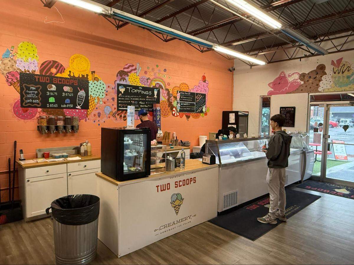 An interior view of an ice cream shop with a bright orange mural wall featuring illustrations of ice cream cones and toppings. A teenager in a grey windbreaker stands at the white service counter waiting for an order. Black menu boards list prices for scoops, pints, and various toppings.