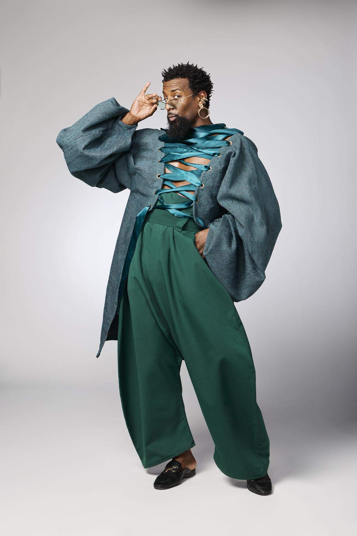 A full-length studio portrait of a man in a high-fashion ensemble. He models an oversized, textured teal coat with voluminous sleeves, laced across his bare chest with a wide, emerald green satin ribbon. He pairs it with matching emerald green, wide-leg trousers, large gold hoop earrings, and black mules. He poses dynamically, adjusting his glasses while looking at the camera.
