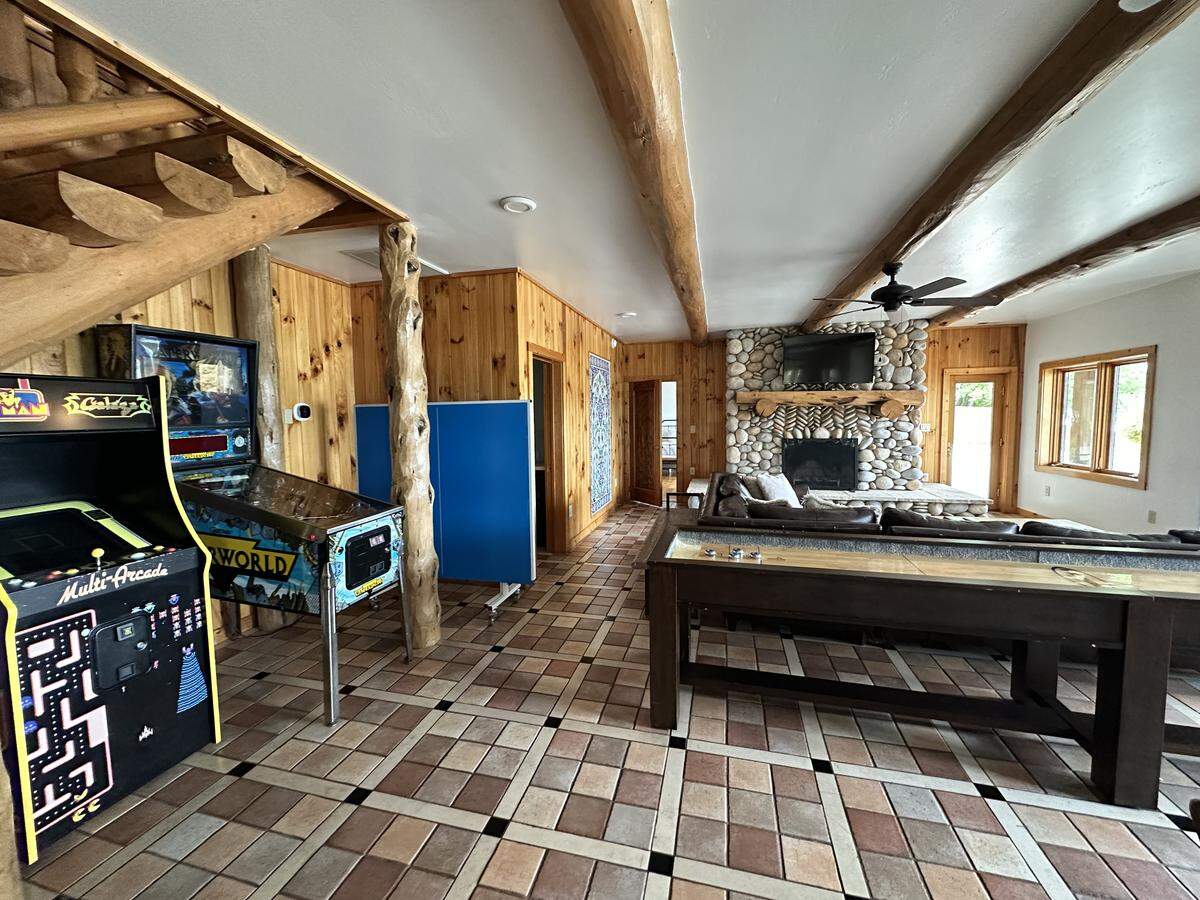 A wide shot of a recreation room in a log cabin. On the left, there is a Pac-Man multi-arcade cabinet, a Centipede arcade machine, and a pinball machine. In the center of the room is a long, wooden shuffleboard table. In the background, a large sofa is arranged around a floor-to-ceiling stone fireplace with a TV mounted above it.