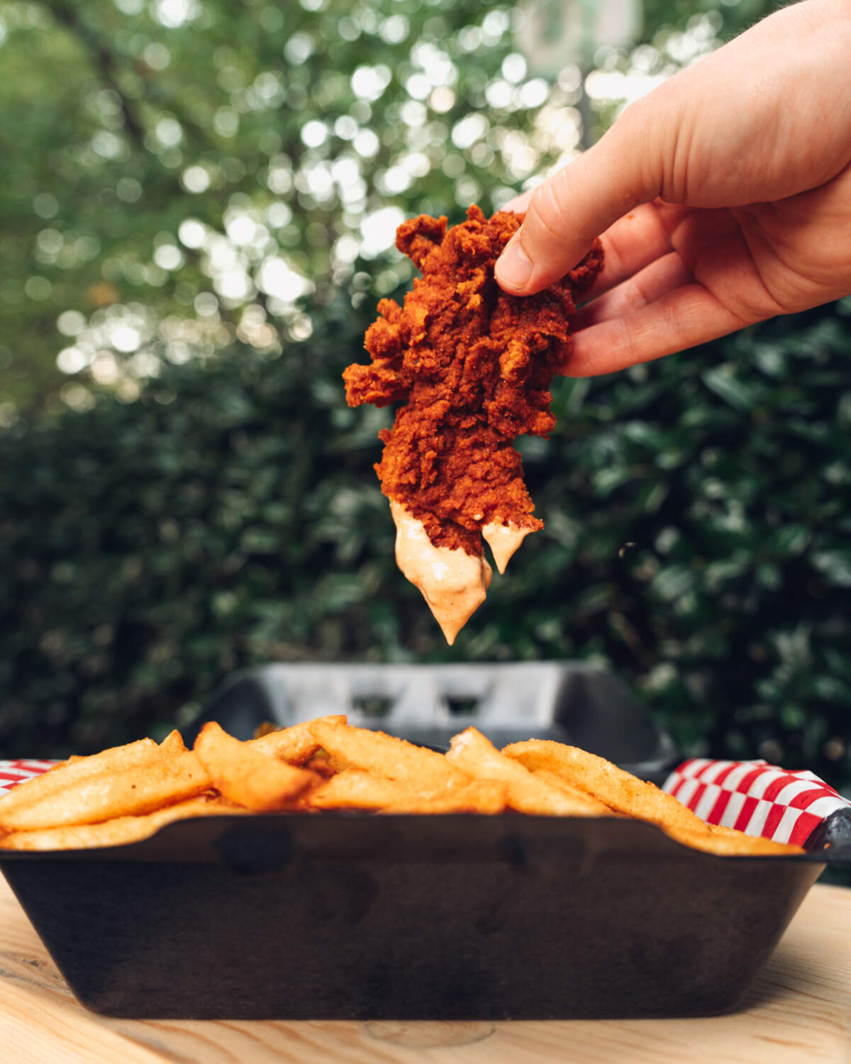 A hand dunks a crispy, red-seasoned chicken tender into a creamy dipping sauce, held above a basket of french fries with a blurred green, outdoor background.