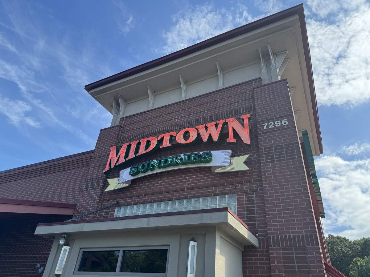 Exterior brick facade of the “Midtown Sundries” restaurant under a bright blue sky with wispy clouds. The building features a prominent sign with “MIDTOWN” in large, red block letters mounted above “SUNDRIES” in green lettering on a stylized white banner graphic. The building number “7296” is visible on the brick column to the right of the sign, and a glass-block window sits directly below it.