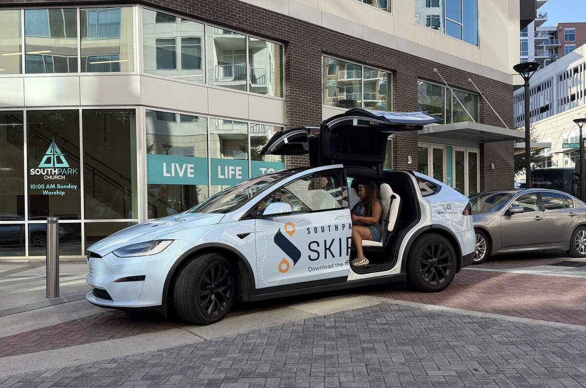 A light blue Tesla Model X with a “SouthPark Skipper” logo wrapped on the side parked outside a building, showing its falcon-wing passenger doors open as a woman sits inside.