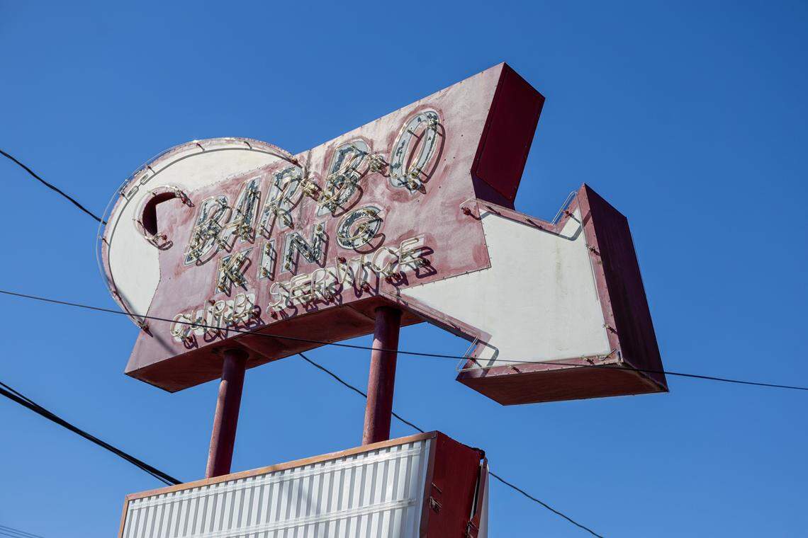 A low-angle shot looking up at the large, weathered, retro sign for “Bar-B-Q King.” The red and white sign, featuring an arrow pointing to the right and exposed neon tubing, stands out against a clear, bright blue sky.