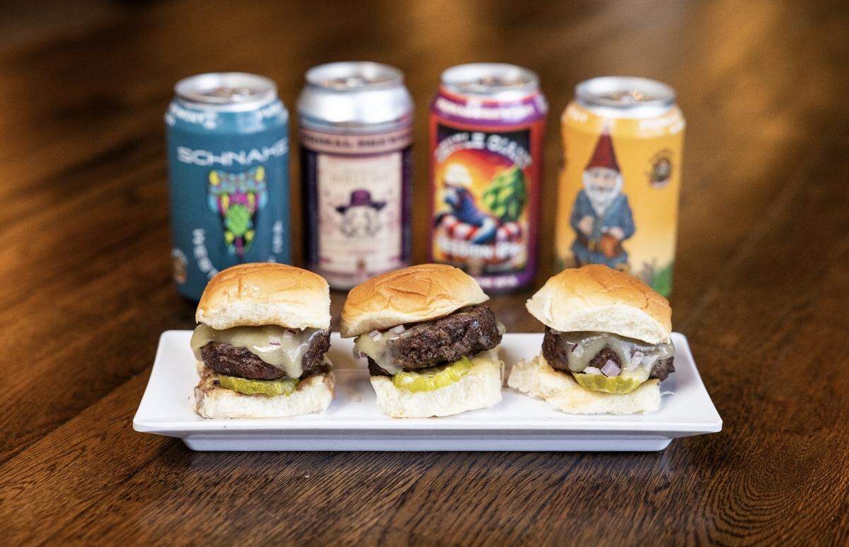 A trio of beef sliders served on a white rectangular plate. Each slider features a thick patty topped with melted cheese, a pickle slice, and red onion on a brioche bun. Behind the sliders, four colorful craft beer cans from Primal Brewery are lined up in a row on a wooden table.