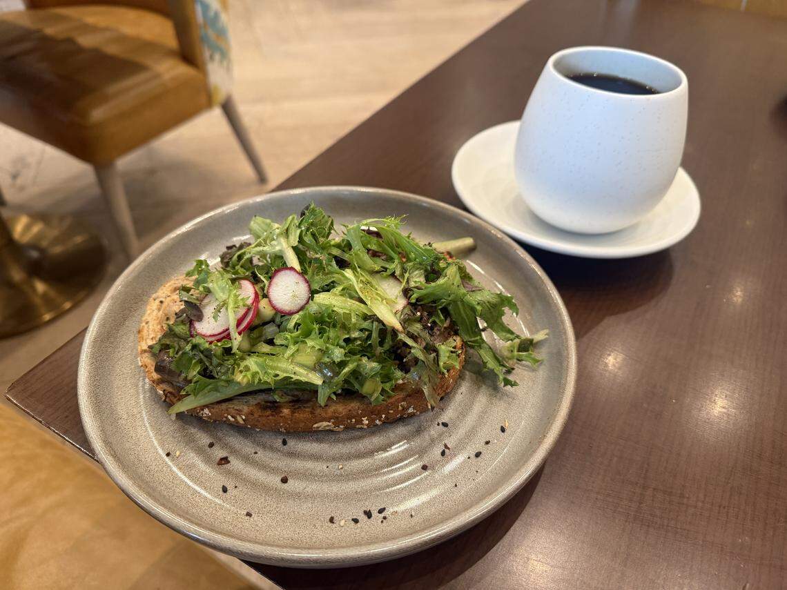 A breakfast or brunch scene featuring a slice of thick, toasted multigrain bread topped with a heap of fresh green frisée salad and thin slices of red radish. The plate is sprinkled with black pepper and seeds. In the background, a white ceramic mug filled with black coffee sits on a matching saucer.