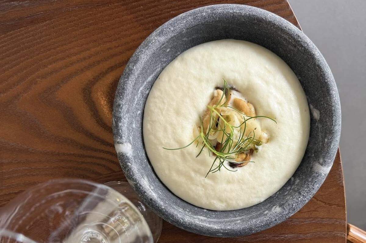 An overhead, close-up shot of a creamy white soup or purée served in a textured, dark gray stone bowl. The center of the dish is garnished with sautéed shellfish (mussels or clams) and fresh green herbs. The bowl sits on a wooden table next to a wine glass.
