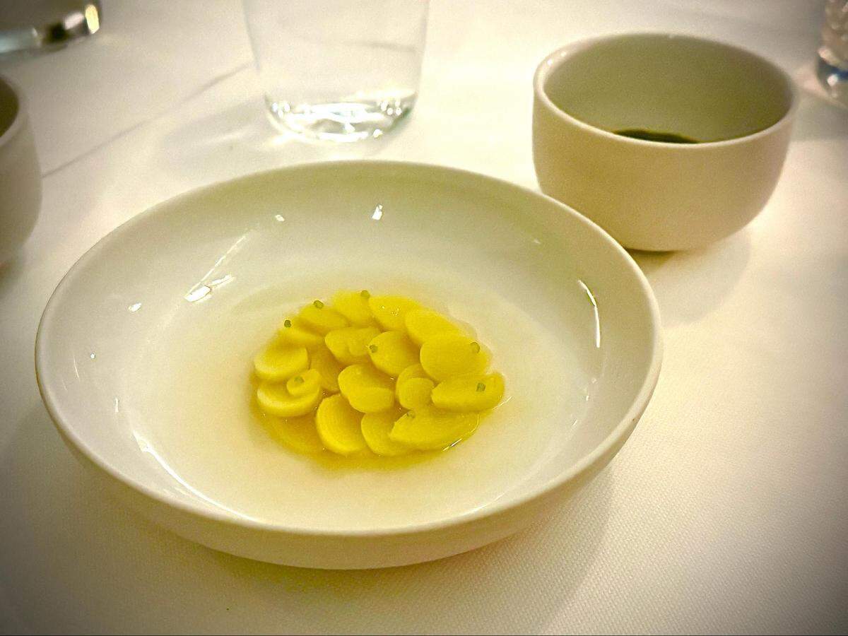 A high-angle, close-up shot of a white, shallow bowl containing a clear broth. In the center, a cluster of small, bright yellow, overlapping discs (possibly pasta or sliced vegetables) is arranged, with tiny green garnishes on top. In the blurred background are a drinking glass and a small, white cup.