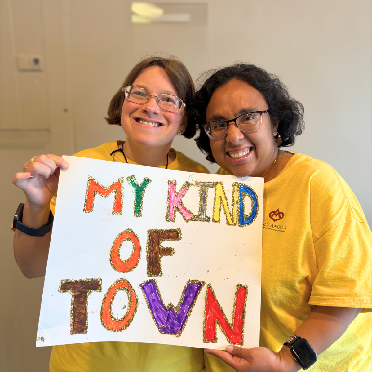 A photo shows two smiling people standing side-by-side, holding a white sign with the colorful, glittery text “MY KIND OF TOWN” written on it. Both people are wearing yellow t-shirts and glasses, and they are looking directly at the camera with happy expressions.