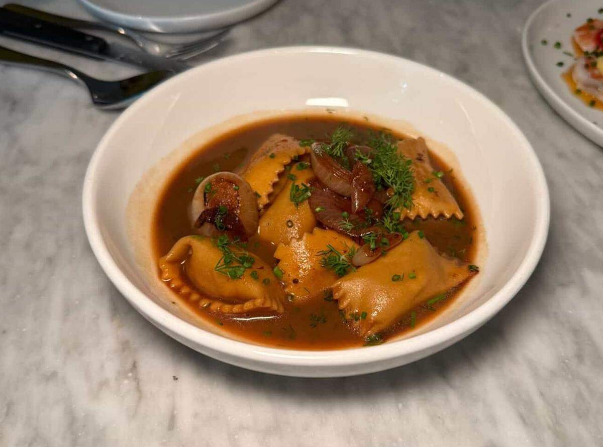 A white bowl on a marble surface holds a dish of “Customshop’s Lamb Agnolotti with onion brodo.” Several plump, golden-brown agnolotti pasta are submerged in a rich, reddish-brown broth. The dish is garnished with tender, halved pearl onions, fresh green herbs like dill and parsley, and finely chopped chives. A fork and knife are visible in the blurred background to the left, and a portion of another plate is seen on the right.