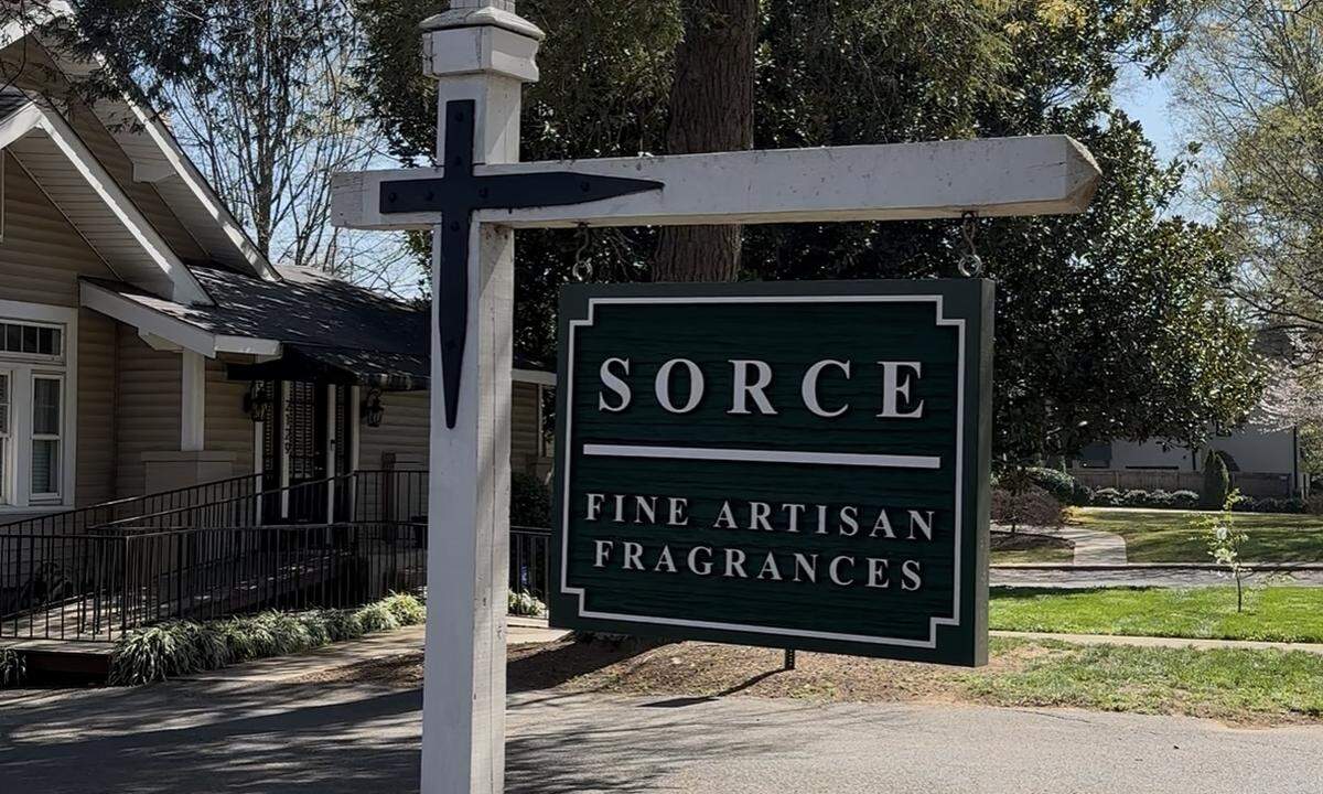 A dark green wooden hanging sign on a white post. The sign reads “SORCE” in large white serif letters, followed by a horizontal line and the words “FINE ARTISAN FRAGRANCES” below it. The background shows a sunny day with green grass and the corner of a residential house.