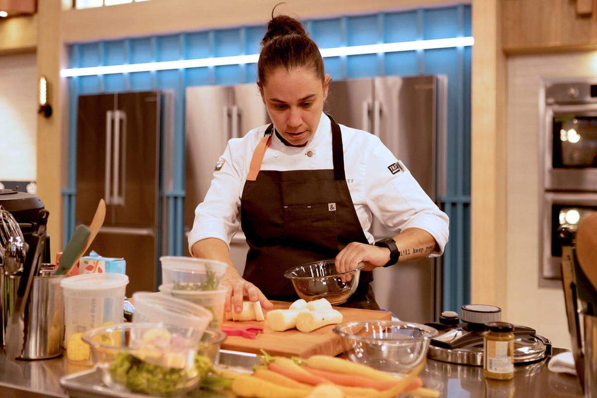 A medium shot of a chef with her hair in a bun, wearing a white chef’s coat and a black apron, focused on food preparation in a professional kitchen. She is working at a stainless steel station with a wooden cutting board, surrounded by clear prep containers, glass bowls, and fresh vegetables like carrots and parsnips. The background features modern blue wall paneling, industrial refrigerators, and warm lighting.