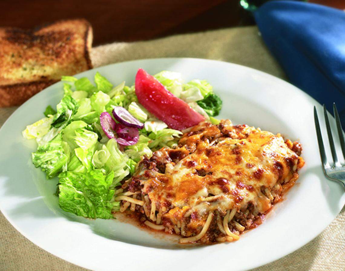 A white plate holding a serving of baked spaghetti topped with meat sauce and melted cheese. It is served alongside a green salad with red onion and a slice of red tomato or pickled relish, with a piece of toasted garlic bread and a fork visible on the side.