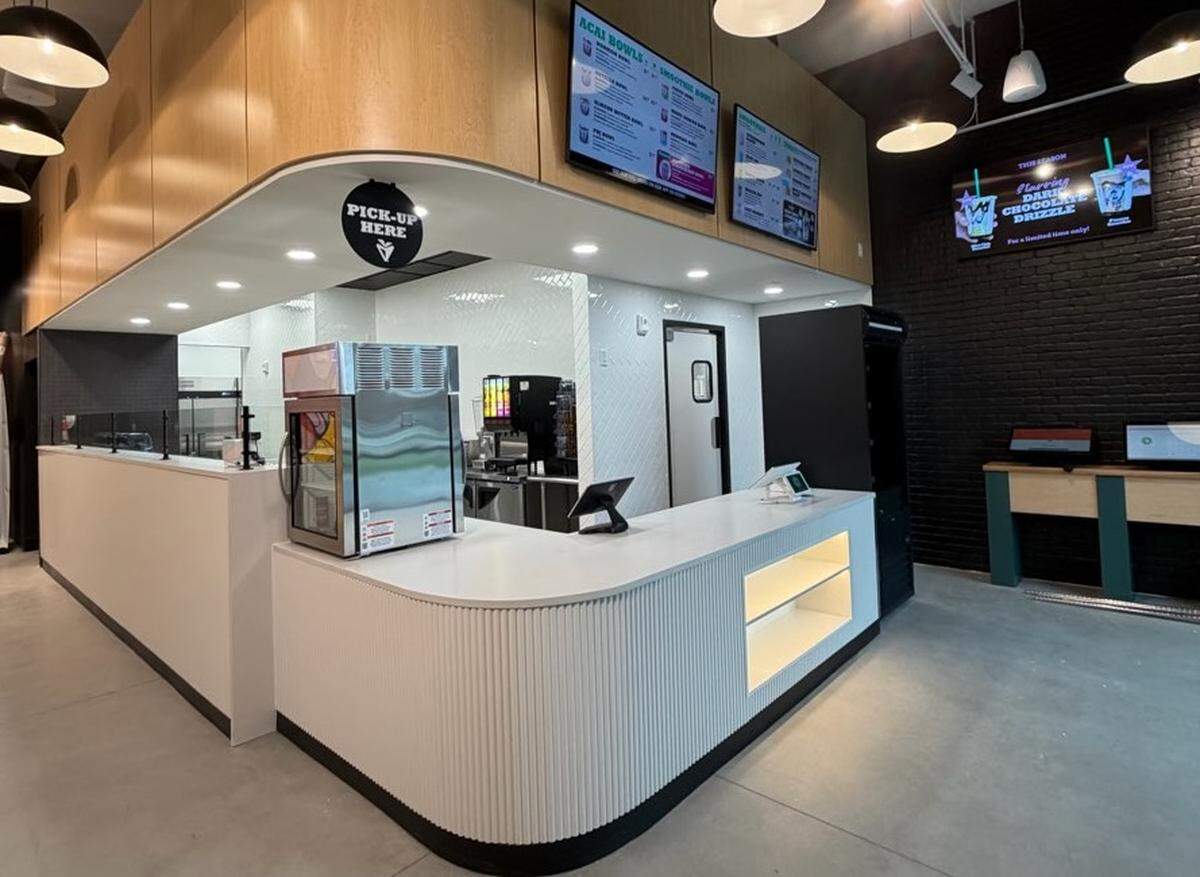 The modern interior of a South Block location, featuring a white ribbed service counter with a white countertop. Two digital menu boards are mounted on a light wood bulkhead above the counter. To the right is a black brick wall with two self-service kiosks and a screen displaying a “Dark Chocolate Drizzle” promotion.