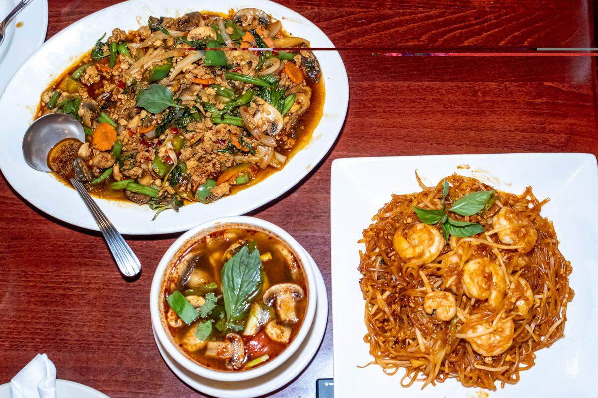 An overhead view of a full meal spread on a wooden table, including a large plate of shrimp Pad Thai, a bowl of Tom Yum soup with mushrooms and herbs, and a plate of basil stir-fry.