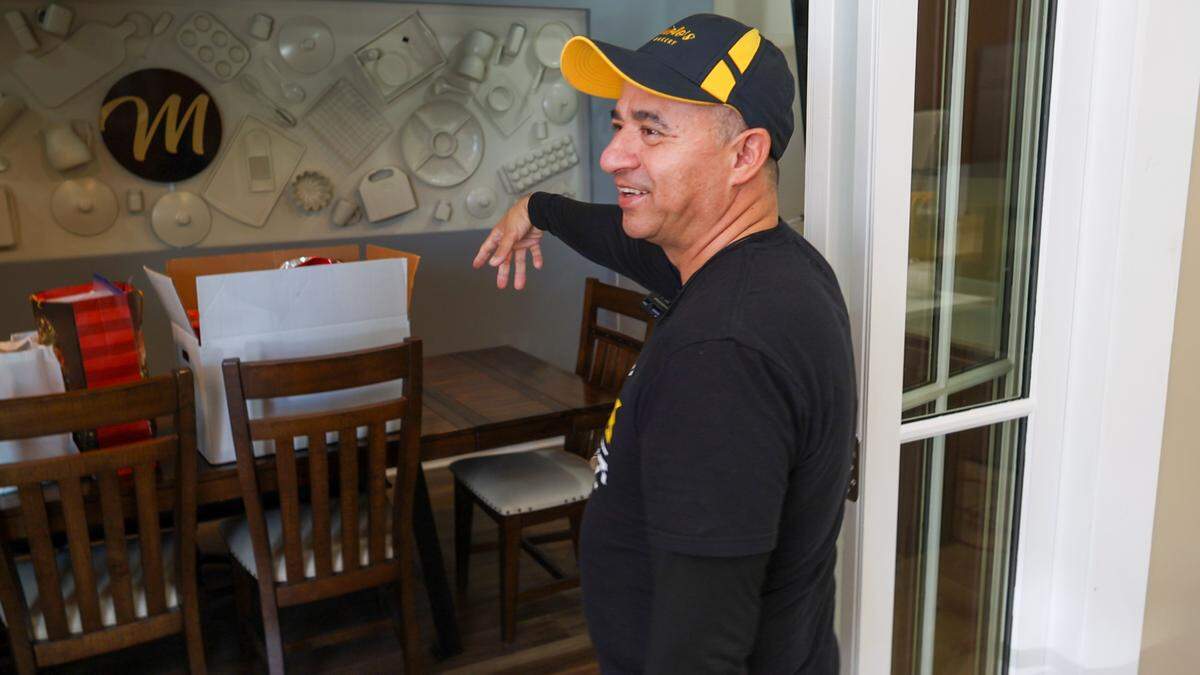 A person wearing a black t-shirt and a black and yellow baseball cap stands in a doorway, gesturing toward a wooden dining table. The wall behind the table features a large circular logo surrounded by various white kitchen-themed wall decorations.